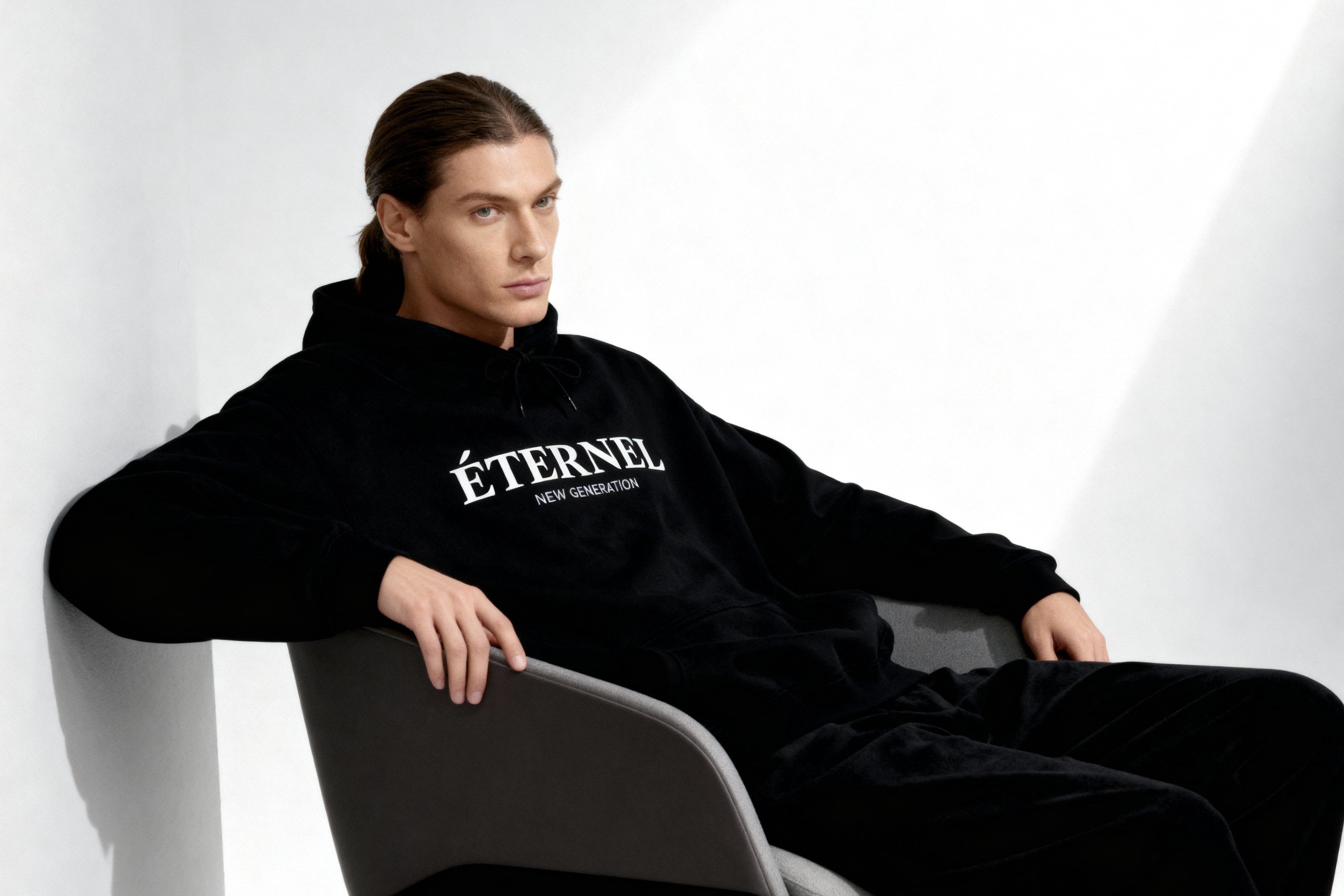 Person wearing a black 'ÉTÉNEL' hoodie sitting on a chair against a white background